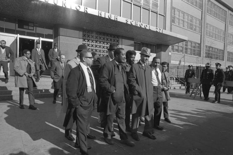 Un groupe d’hommes noirs se rassemblent devant un bâtiment scolaire.