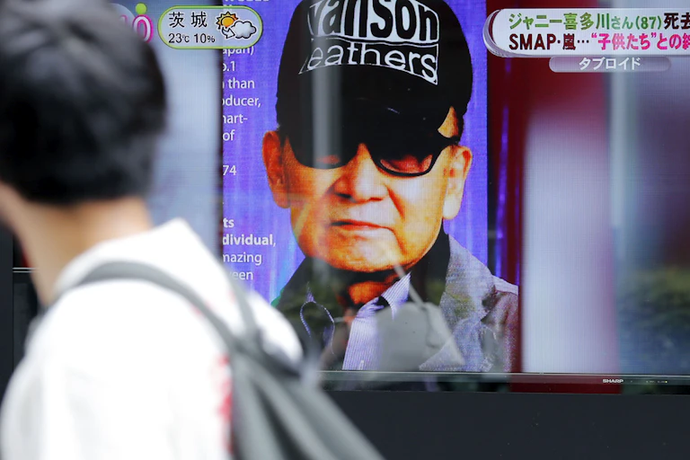 A man in a black ballcap and dark glasses seen on a screen as another man walks past.