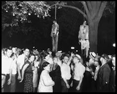 A black and white photograph of a large crowd of white people looking up, many of them grinning, at tree branches where two men have been hanged, their bodies dangling from the branches.