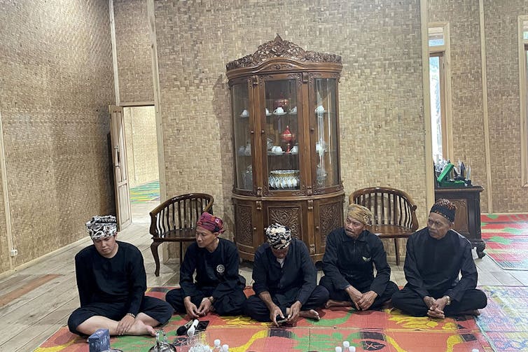 A group of five Indonesian men sit on a rug in a sparse but elegant room with a hutch in back. This is a room where they receive visitors. The men are royalty in this Indonesian community