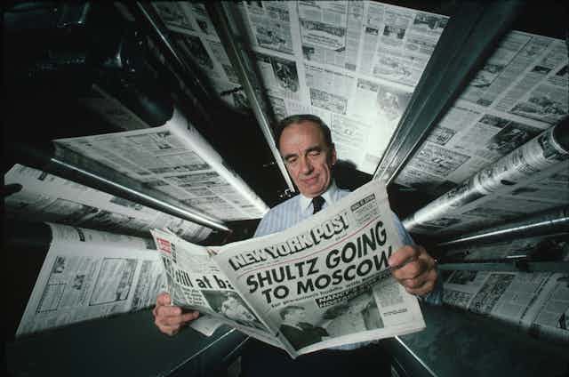 A man in shirt and tie reads a newspaper as printing presses show pages above his head.