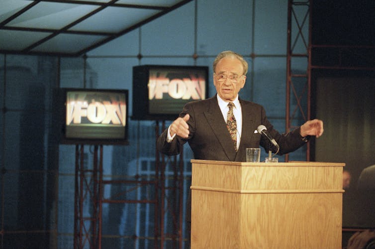 A man gesture while talking at a podium, behind him TV screens show 'Fox'