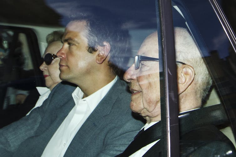 Two men in suits are seen through the back window of a car.