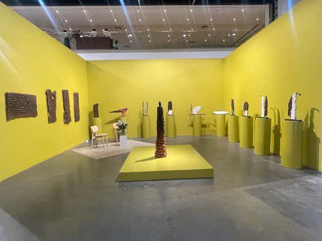 A gallery space with yellow walls and plinths, which house 10 sculptures in brown and white, four wall hangings and a large conical sculpture in the centre.