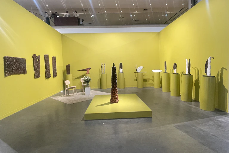 A gallery space with yellow walls and plinths, which house 10 sculptures in brown and white, four wall hangings and a large conical sculpture in the centre.