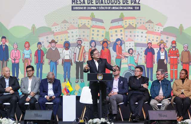 Seis homens e uma mulher sentados em cadeiras, num palanque, ouvem o presidente colombiano, que discursa em pé.