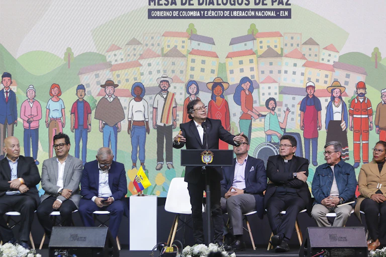 Seis homens e uma mulher sentados em cadeiras, num palanque, ouvem o presidente colombiano, que discursa em pé.