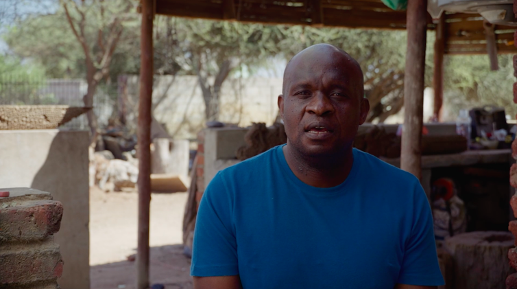 A man in a blue T-shirt with a bald head sits outside, in front of a wooden structure and thorn trees.