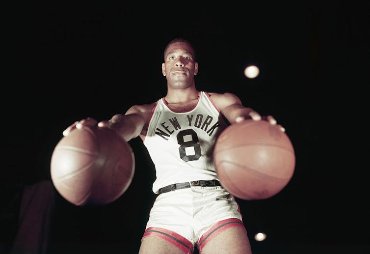 Young Black man in white basketball jersey palming a basketball in each hand.