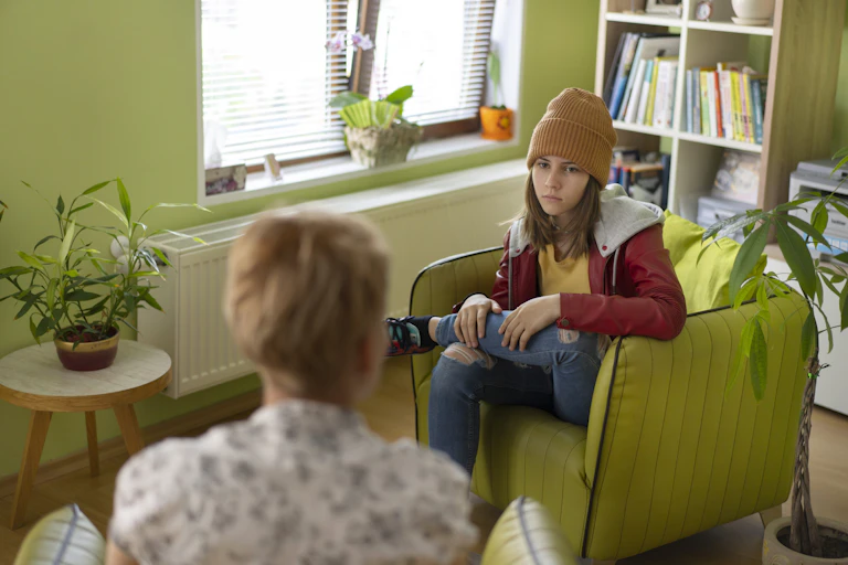A teen girl in jeans, a hoodie and beanie cap sits, looking serious, in a green chair in a green office with a woman with blonde hair.