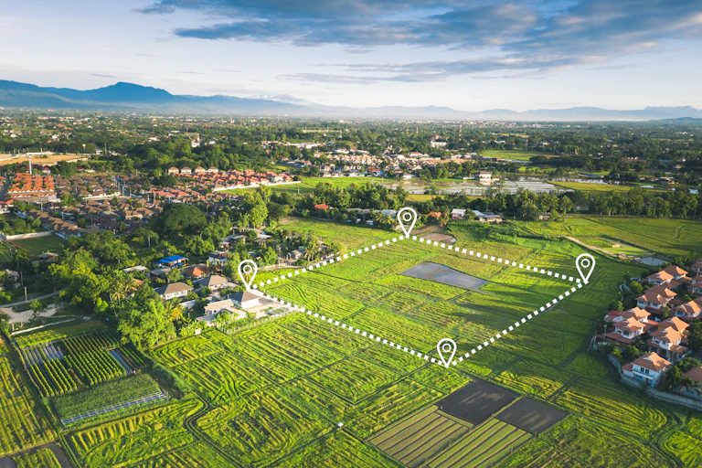 Aerial view of green fields with digital map pins marking out a plot, some housing developments nearby, mountains and sky in the background.