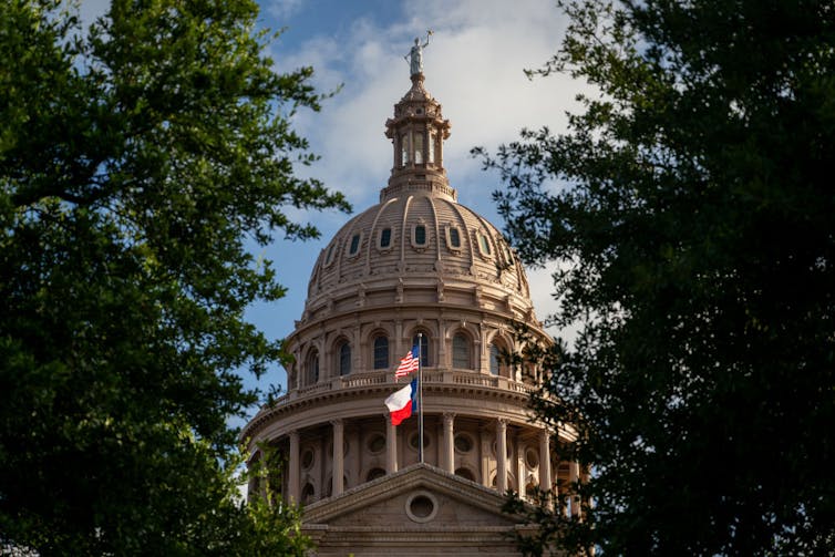 The multistory dome of a large building is seen between trees, with two flags flying in front.