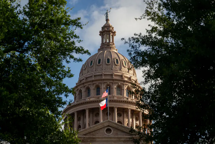 The multistory dome of a large building is seen between trees, with two flags flying in front.