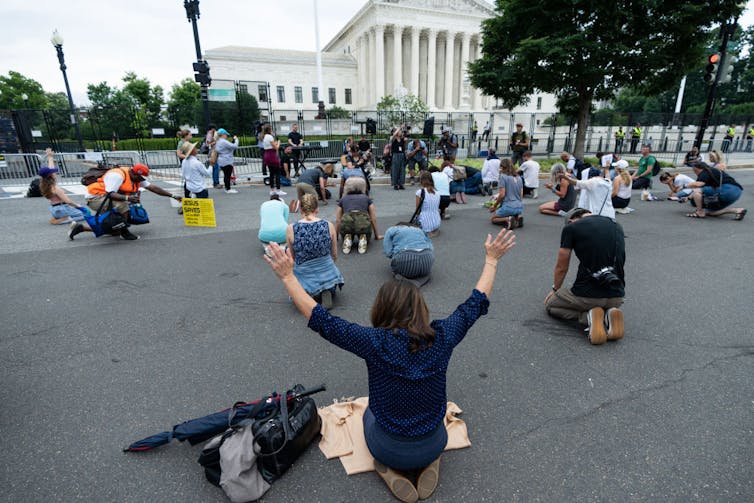 People kneel on a large piece of pavement, looking toward a large white building with columns.