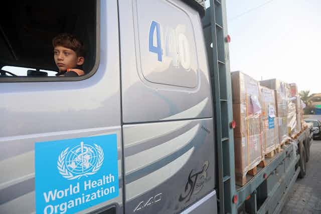 A young boy sits in the front seat of a silver truck that has the World Health Organization logo o it. Behind the truck large boxes are seen.
