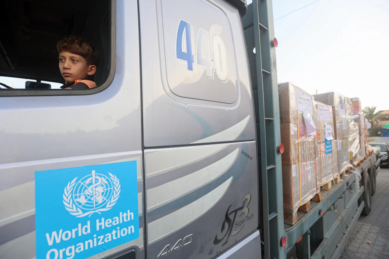 A young boy sits in the front seat of a silver truck that has the World Health Organization logo o it. Behind the truck large boxes are seen.