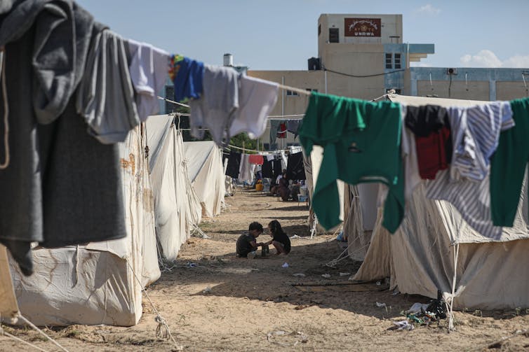 Deux enfants sont assis par terre entre des rangées de tentes blanches et des vêtements accrochés à des cordes à linge blanches.