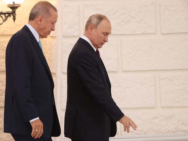 Two men wearing dark suits chat as they walk past a brick wall.