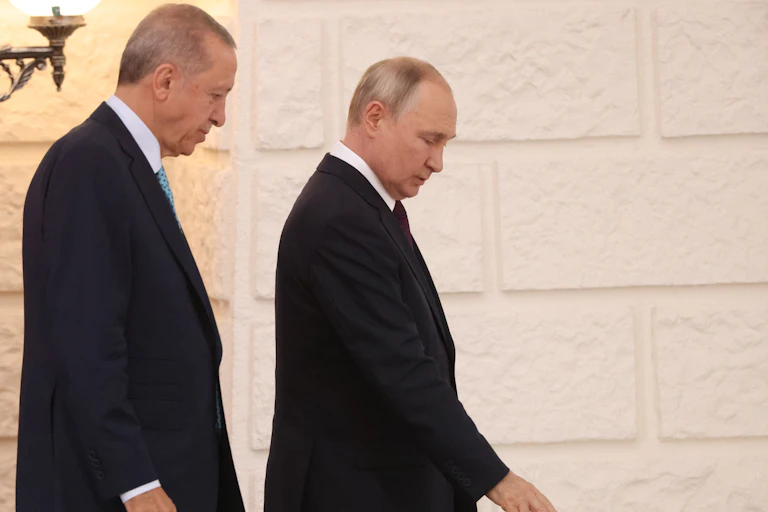 Two men wearing dark suits chat as they walk past a brick wall.