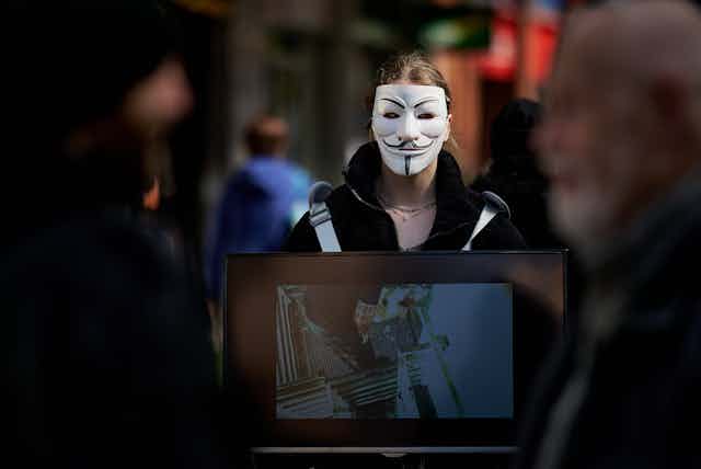 A man in an anonymous mask holding a computer monitor outdoors.
