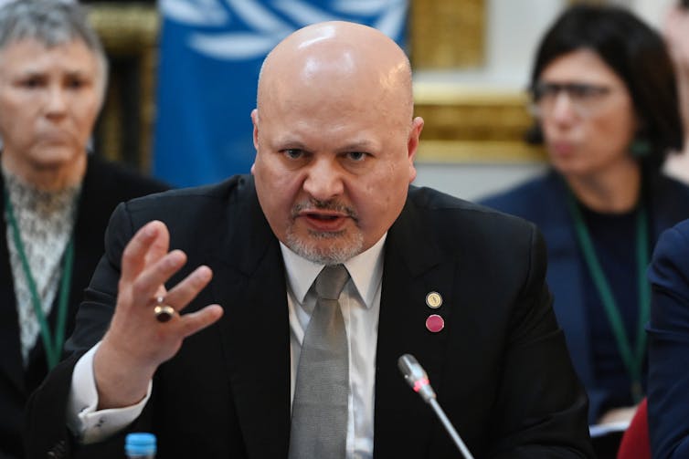A photo showing a bald man in a suit holding forth in front of a microphone with two out-of-focus people in the background.