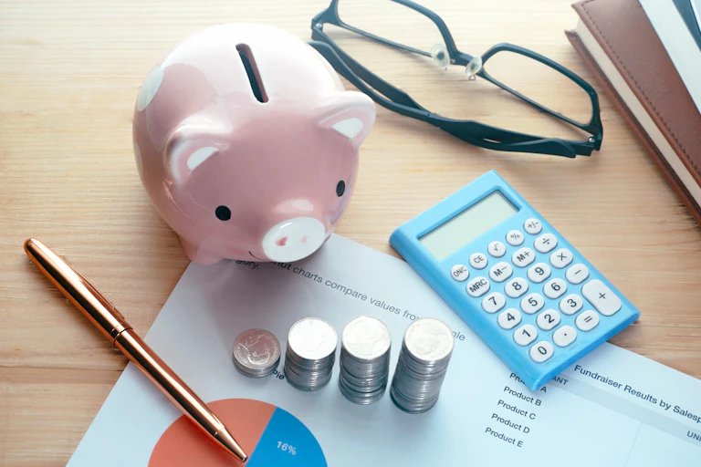A piggy bank, a calculator, a pen and several stacks of coins sitting on a desk