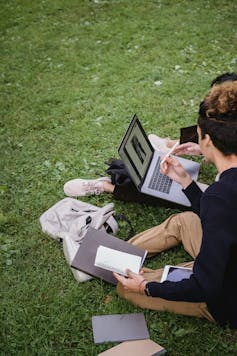 People sit on a grassy lawn surrounded by bags, books and laptops.