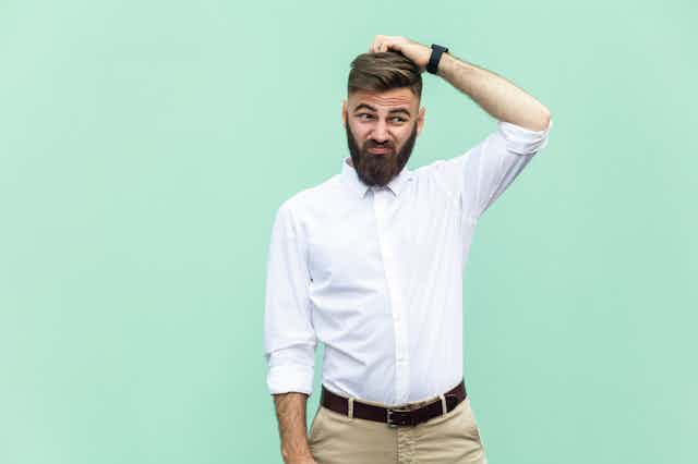 A young man wearing a dress shirt and trousers looks confused and scratches his head in front of a blank, mint green background