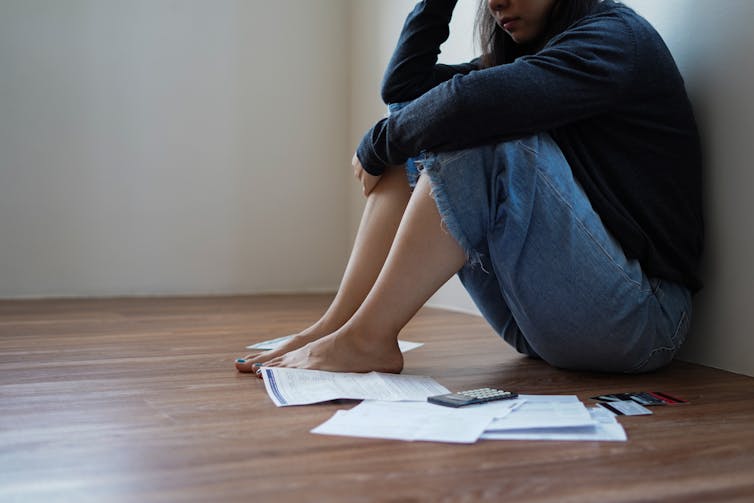 A woman sits on the floor resting her head in her hands, with financial papers around her feet
