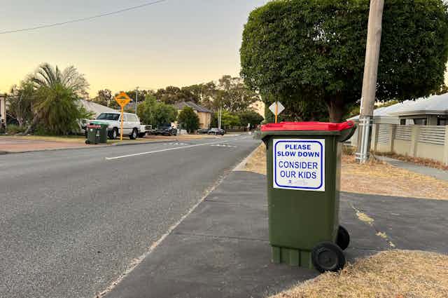 A waste bin at the side of a residential road, with a sticker on the side saying "Please slow down. Consider our kids."
