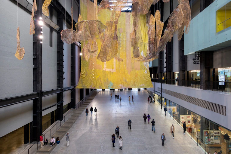 A large art gallery space, people dwarfed by two giant works, one a shimmering cloth, the other the shape of the continents on earth.