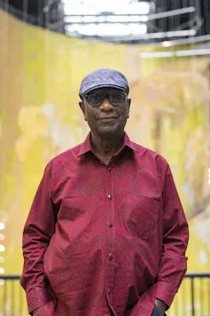 An elderly man in glasses, a beret and a red shirt stands with a wry smile in front of a large, golden, hanging artwork.
