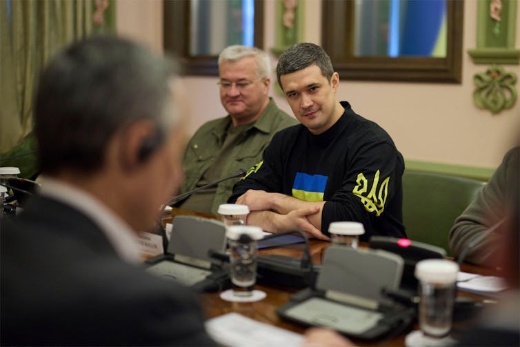 Three men in a room, one is wearing a black sweatshirt with the Ukraine flag in it.