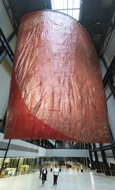 A vast red sculpture hanging from the roof, people walking below it.