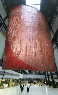 A vast red sculpture hanging from the roof, people walking below it.