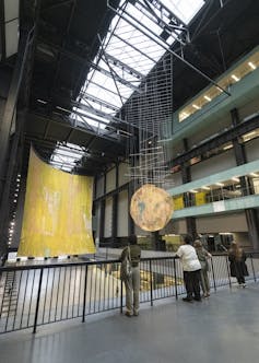A large yellow sculpture shimmers with a earth-like sculpture hanging in front of it.
