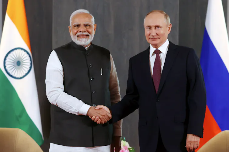 Two men shake hands, one in a suit and tie, with colourful flags behind them.