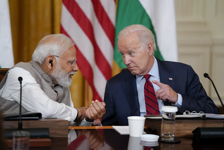 An older man in a suit and tie raises his finger while talking to a grey-haired man who leans towards him.