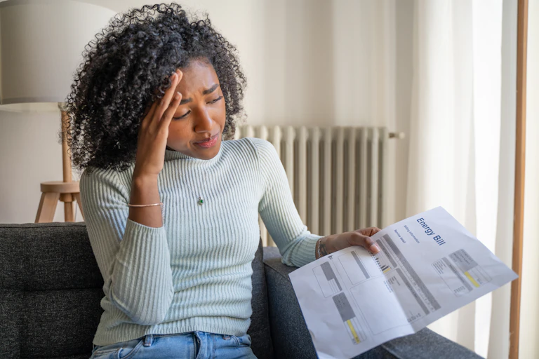 Woman holding energy bill, looking worried, sitting in living room.
