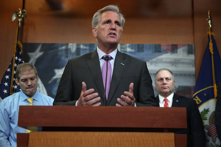 A middle aged white dressed in a business suit rests his hands on a lectern as he stands in front of two other white men