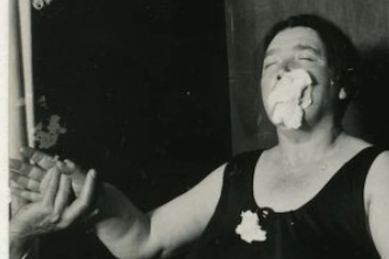 A woman seen holding hands with people on either side of her and a floating white mass around her mouth.