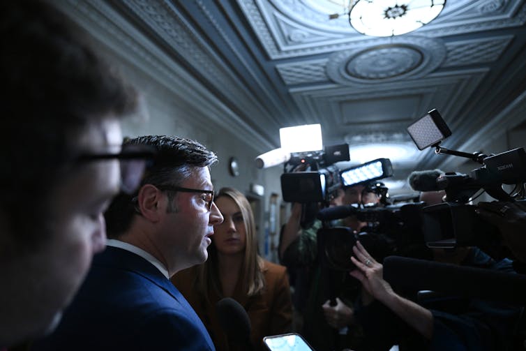 A middle aged white man wearing eyeglasses is surrounded by television cameras and reporters.