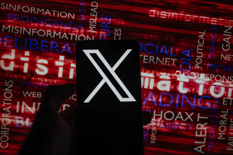 X social media icon logo displayed on a smartphone with words like disinformation seen on screen in the background.