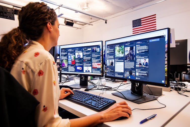 A woman sits facing two computer screens while scrolling through news stories.