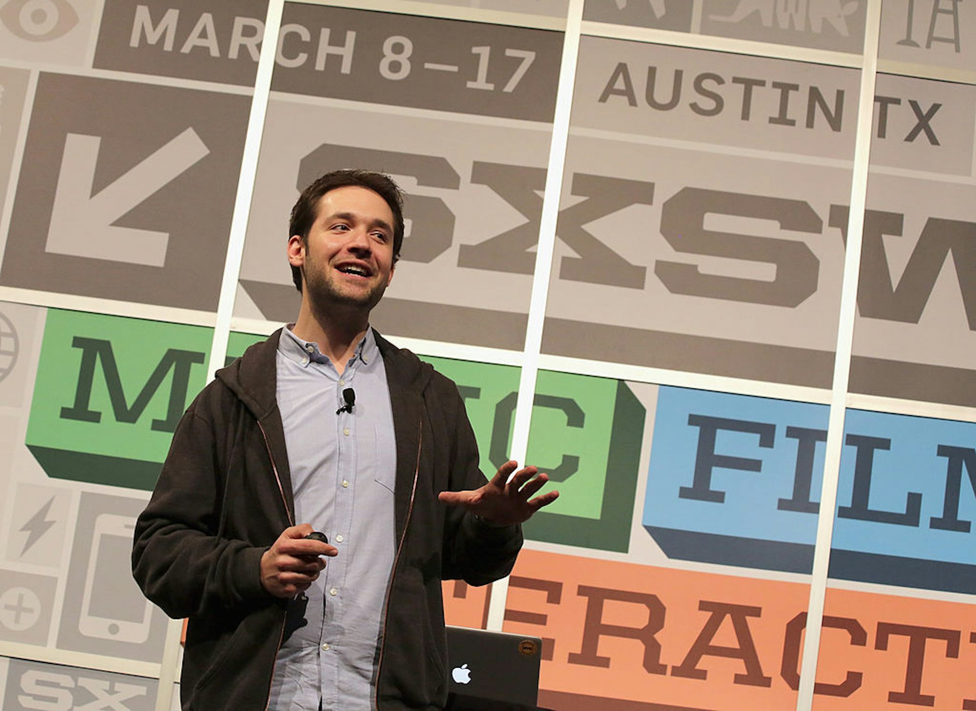 Man wearing zip-up sweatshirt speaks on stage.