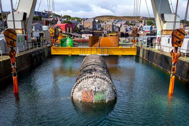A photo shows a large, barnacle-encrusted cylinder floating in the sea inside a metal gantry.