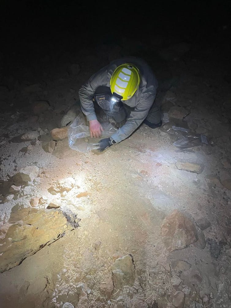 A photo of a man wearing a helmet with a torch, crouching in a dark cave and inspecting the ground.