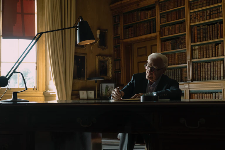 Man sits at writing desk with bookshelves behind him