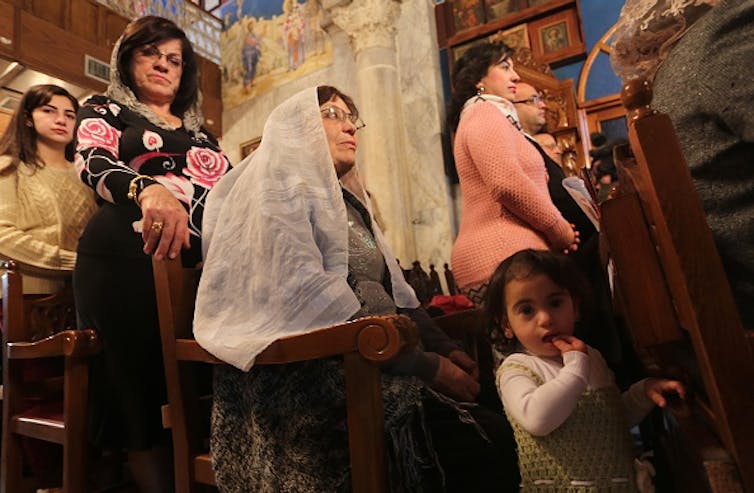 Women, with their head covered, stand in church pews, along with children.