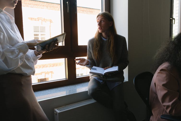 A seated man with an open notebook talks to a woman who is standing holding a tablet.